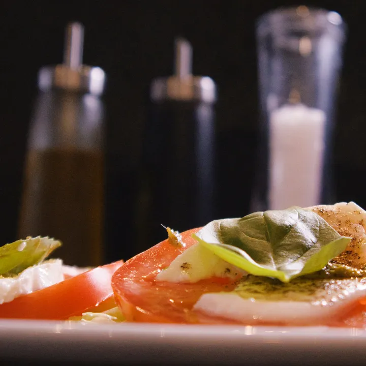 Caprese auf weißem Teller: abwechselnd Tomatenscheiben und Mozzarella mit Basilikumblättern und Olivenöl, dahinter unscharf Öl- und Essigkaraffen sowie Salz und Pfeffer.
