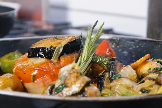 Schwarze Pfanne auf dem Herd in der Küche, darin gebratene Aubergine, Tomate, Zucchini und Kartoffeln, obenauf ein Zweig Rosmarin.