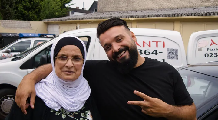 Der Inhaber im schwarzen T-Shirt und seine Mutter mit hellem Kopftuch stehen lachend Arm in Arm im Hof hinter der Pizzeria, dahinter die geparkten Lieferfahrzeuge.