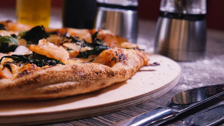 Nahaufnahme des aufgegangenen, leicht angebrannten Pizzarandes auf einem hölzernen Servierbrett, im Hintergrund zwei Pfeffermühlen und eine weinrote Wand.