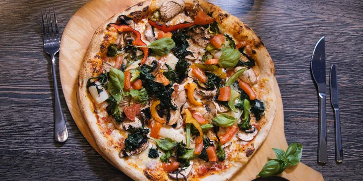 Gemüsepizza von oben, dicht belegt mit gegrillter Paprika, Zucchini, Champignons, Stängelkohl und Tomatenstücken, daneben Messer und Gabel auf dunklem Holztisch.
