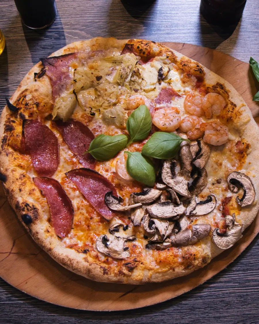 Pizza Quattro Stagioni von oben auf einem hellen Holzbrett: je ein Viertel mit Kochschinken und Artischocken, Garnelen, Salami und Champignons, in der Mitte frisches Basilikum.