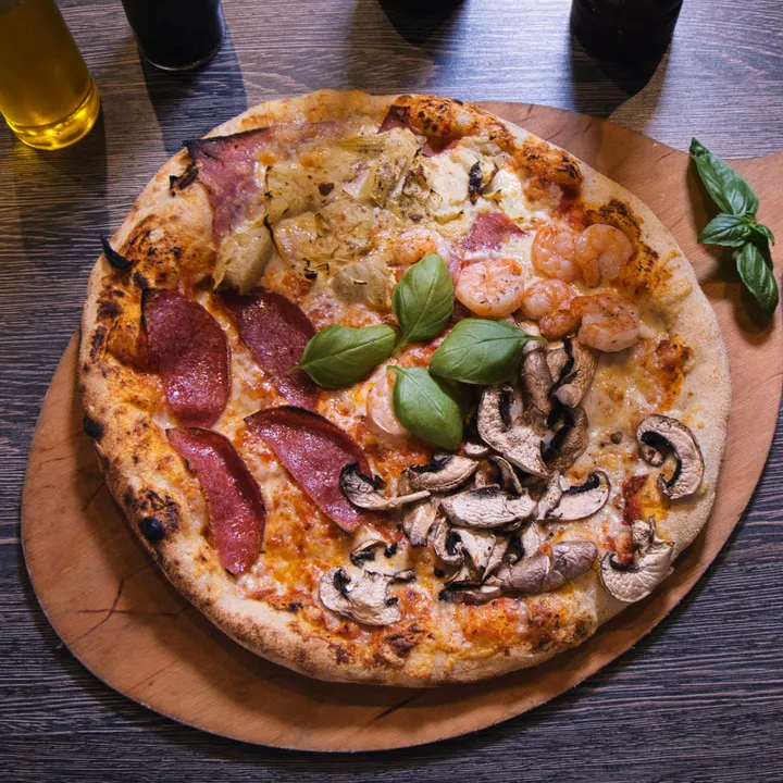 Pizza Quattro Stagioni von oben auf einem hellen Holzbrett, in vier Viertel belegt mit Schinken und Artischocken, Garnelen, Salami und Champignons, in der Mitte Basilikum.